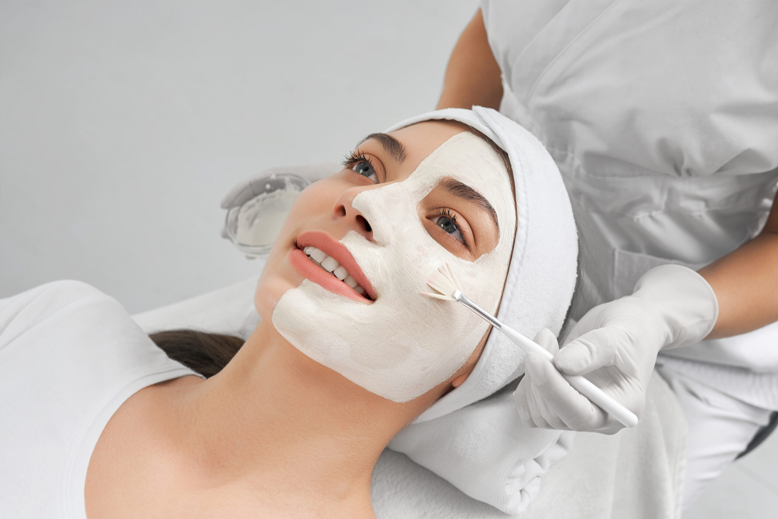 Side view portrait of beautician in rubber gloves and white uniform doing special mask for young smiling woman in beauty salon. Concept of process procedure for skin rejuvenation.